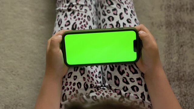 A little girl is sitting at home on rug wearing leopard-print leggings, holding horizontaly new smartphone in her hands. Phone is dressed in a green case, and on the screen, there is a chroma key.