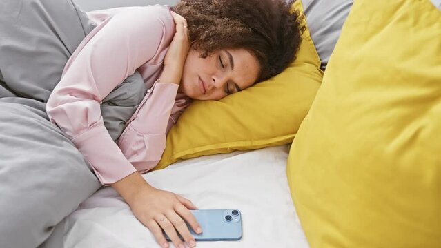 Young hispanic woman with curly hair sleeping in bed, wearing pajamas, in cozy bedroom setting.