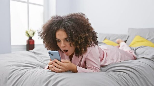 Astounded hispanic woman with curly hair lies on bedroom bed, in profound shock from smartphone surprise. disbelief paints her face, mouth agape in wonder, revealing a scared yet amazed expression.