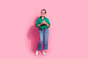 Fototapeta premium Full body photo of attractive pensioner woman hold telephone look empty space wear trendy green clothes isolated on pink color background