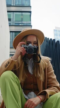 Vertical shot stylish female street photographer in beige hat and coat posing outdoors in modern city. Beautiful blonde woman sitting on stairs smiles holding retro style photo camera in her hand
