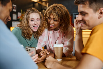 excited woman showing smartphone to happy multiethnic mates in lounge cafe of students hostel