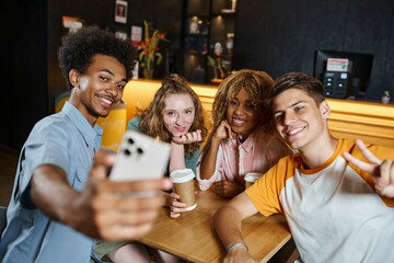 Fototapeta premium cheerful african american man taking selfie with stylish multiethnic friends in lounge of hostel