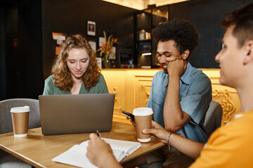 young smiling woman using laptop near multiethnic men with smartphone and notebook in hostel cafe