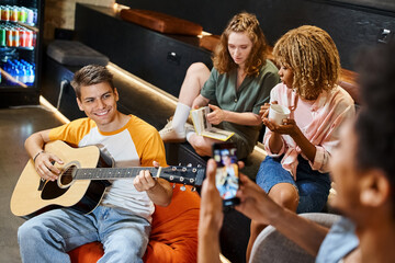 african american man taking photo of cheerful friend playing guitar in lounge of students hotel