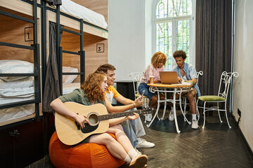 multicultural students with guitar and laptop in room of contemporary youth hostel, travelers - obrazy, fototapety, plakaty
