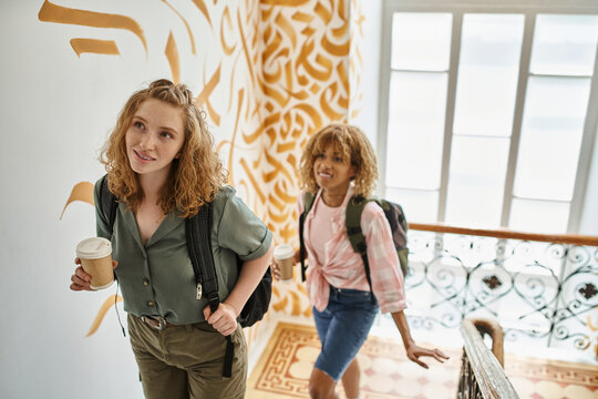 youthful multiethnic female students with paper cups and backpacks walking on hostel staircase