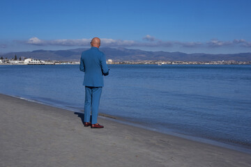 Elegant gentleman in a blue suit, strolling on Poetto Beach: a stylish contrast of formal attire against the serene coastal backdrop. - 707130776