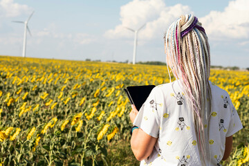 A young woman with a tablet in a field with sunflowers, wind turbines for green energy production,...