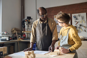 Portrait of Black senior carpenter teaching little boy woodworking in workshop and looking at plans