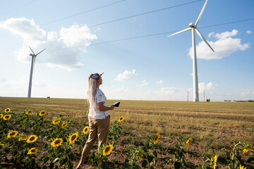 young woman with tablet in field with sunflowers studies wind turbines for production of green...