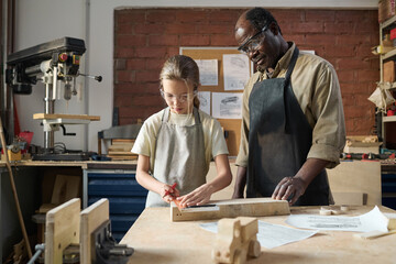 Waist up portrait of young girl measuring wood in workshop with senior carpenter watching