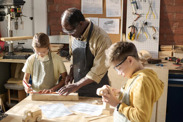 Portrait of senior craftsman teaching children carpentry in workshop lit by sunlight, copy space - obrazy, fototapety, plakaty