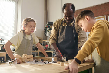 Waist up shot of male carpenter with two children building wooden toys together in crafting workshop