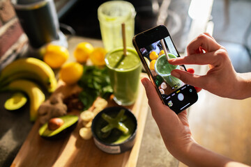 Woman taking photo of a green smoothie with fresh ingredients on kitchen counter