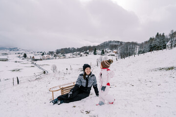 Little girl walks along a snowy hill towards her sitting smiling mother with a sleigh turned on its...