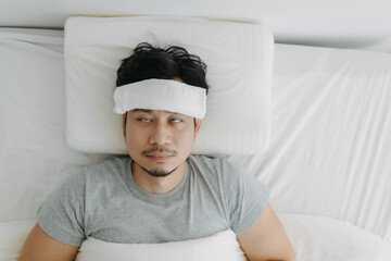 Top view of Asian man got sick with towel on forehead, man flu, man symptoms, resting alone on white bed in apartment, bulge and high fever.