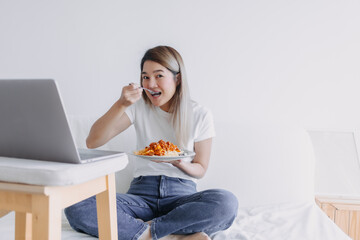 Asian woman eating pasta while watching series from notebook, sitting on white bed and working at home looking at camera.