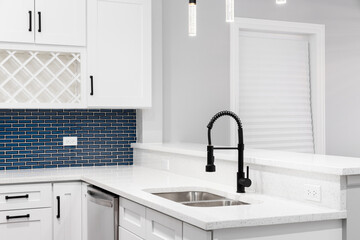 A kitchen detail with a black faucet, glass light fixtures hanging above the white quartz countertops, and a blue glass subway tile backsplash.