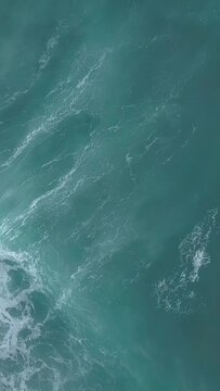 Vertical aerial top view of turquoise sea waves foaming and splashing, big waves from above rolling and breaking on empty ocean beach on cloudy day
