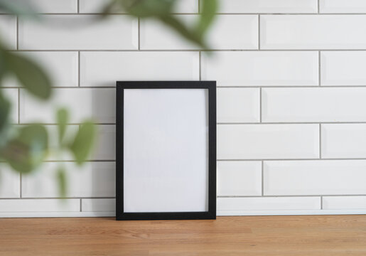 A black frame with a blank canvas against a white tile wall and on a wooden tabletop with blurred green foliage.