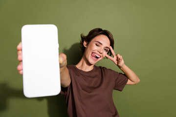 Photo portrait of lovely young lady show v-sign winking hold telephone screen wear trendy brown garment isolated on khaki color background