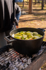 Black pot of white mulled wine cooking on a grill over embers in a forest setting, with a person stirring