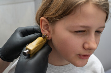 A doctor wearing sterile black medical gloves pierces the ears of a young pretty girl. Girls get their ears pierced. The process of piercing a child's ears in a beauty salon with a piercing gun