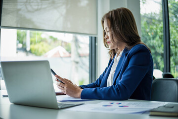 Businesswoman sitting at desk on couch in workplace or at home working on laptop and analyzing data on charts and graphs and writing on papers to make business plan and strategies for company, 