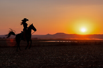 silhouette cowgirl in the sunset riding in the prairie near a waterhole - obrazy, fototapety, plakaty