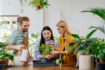 Busy working day at flower shop. Tree coworkers, asian woman, young man and middle age successful blond female owner together. Male watering plant.