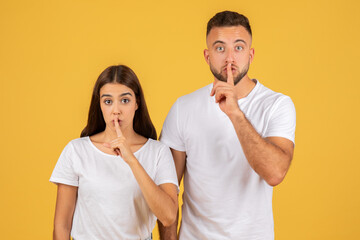 Surprised young couple in white t-shirts with fingers on lips making a shush gesture