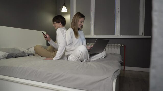 Cheerful people are sitting back to back on a bed, spending time together in a spacious apartment on weekends. A joyful family is browsing social media on wireless gadgets.