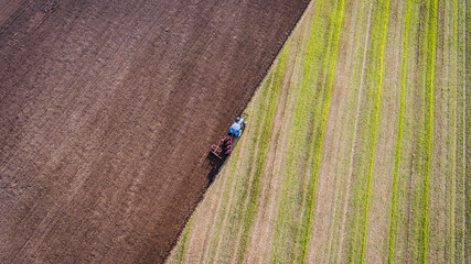 Drone photo of a blue tractor plowing the field. Half of the field on the diagonal is plowed and the soil is brown, the other half is still greenish. The tractor is in between. © Anelia