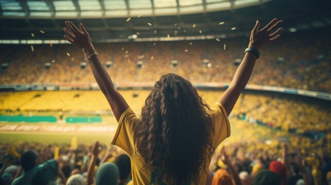 Female soccer fans in yellow Brazilian shirts at the stadium cheer and raise both hands celebrating victory