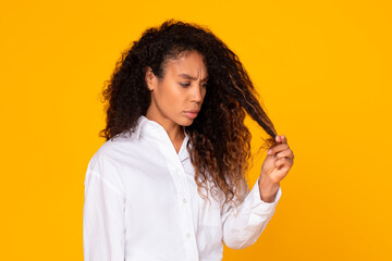 concerned african woman looking at damaged hair ends, yellow background