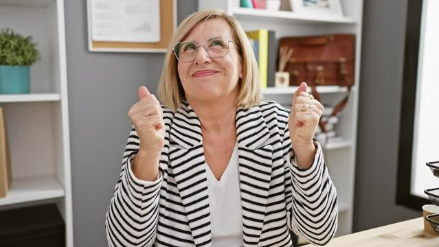 Radiant middle-aged blonde business woman ecstatically celebrates a victorious win, her smile screams triumph, her expression oozes joy in the office setting.