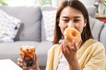 young asian woman enjoy eating sweet donut and drink cola soft drink in living room at home in...