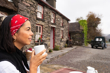 Beautiful latin woman holding cup of coffee while resting next to beautiful stone cottage in the...