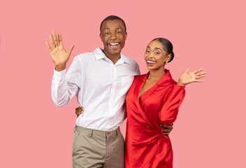 Cheerful black couple waving hands with bright smiles, pink background