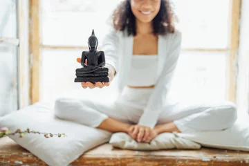 Foto op Aluminium Close up of a Buddha statue on the hand of a dark skinned girl. Young smiling african american woman in white clothes sitting in lotus position on windowsill and holding figurine. © Dasha Petrenko