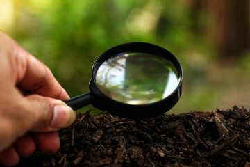 Hand holding a magnifying glass on soil ground with a blurry green background of nature, concept of...