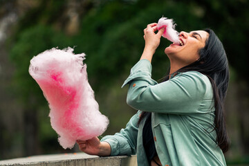 beautiful latin woman sitting, smiling and eating her cotton candy, enjoying the day, in an outdoor...