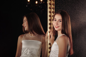 Smiling teen lady in elegant white dress posing at large mirror, happy looking at camera. Portrait of cover teenage girl smile pose in shadow dark room. Youth gen Z emotion concept. Copy ad text space