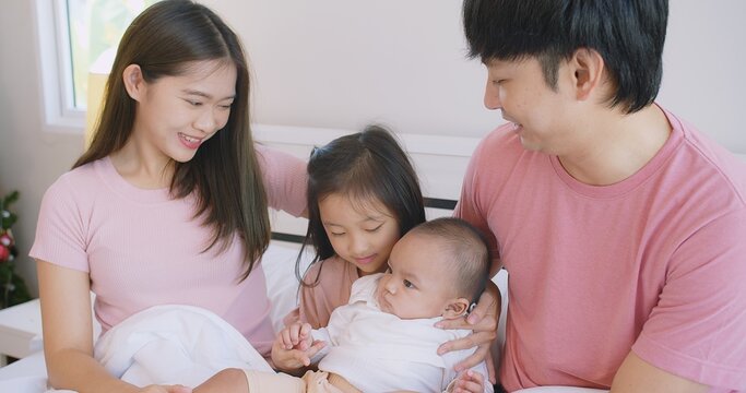 Happy Asian family father mother daughter baby embrace smiling together lying relaxing on white bed in bedroom at home, happy parent and child girl enjoy hugging having fun playing in bedroom