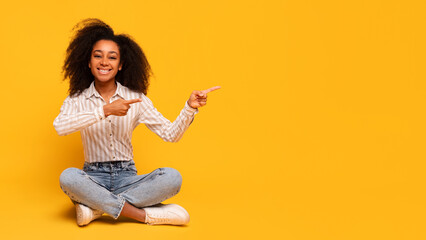 Cheerful black woman sitting cross-legged pointing right at copy space on yellow background