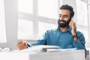 Smiling indian man talking on mobile phone while at office desk - obrazy, fototapety, plakaty