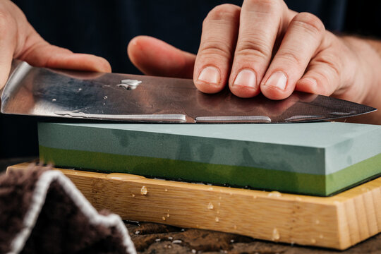 A man sharpens a knife with a grindstone on a rustic wooden table.