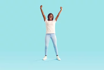 Tuinposter Young woman celebrates success after winning prize. Full body length happy joyful beautiful slim African American girl in casual wear standing on bright blue background, raising hands up and cheering © Studio Romantic