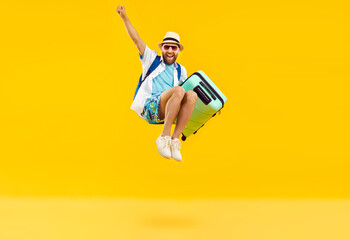 Full body photo of a funny young happy man in sunglasses carrying his blue suitcase jumping and having fun on a yellow background. Summer holidays, vacation trip and travel concept.